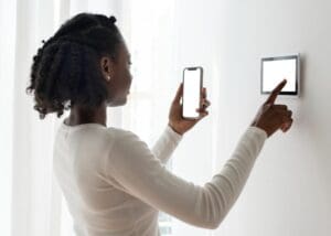 A person in a white long-sleeve shirt uses a smartphone to interact with a wall-mounted smart thermostat.