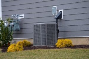An outdoor HVAC unit beside a house wall, with a gas meter and small yellow shrubs in the foreground, set against gray siding.