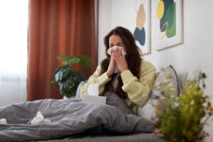 A woman sits on a couch, sneezing from allergies. She has plants on either side of her.